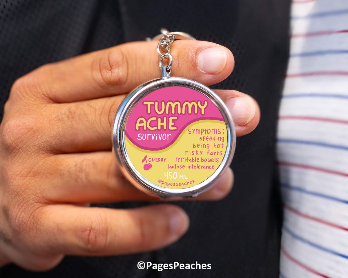 a person holding a keychain with a round label that says &quot;TUMMY ACHE SURVIVOR&quot;.