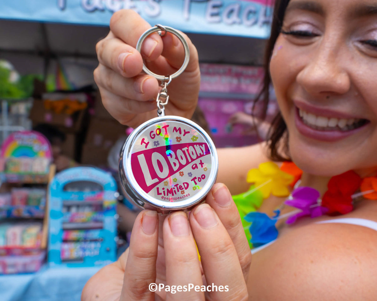 A woman is holding a round keychain with the text &quot;I got my LOBOTY limited edition&quot; on it, smiling and wearing colorful lei garland.