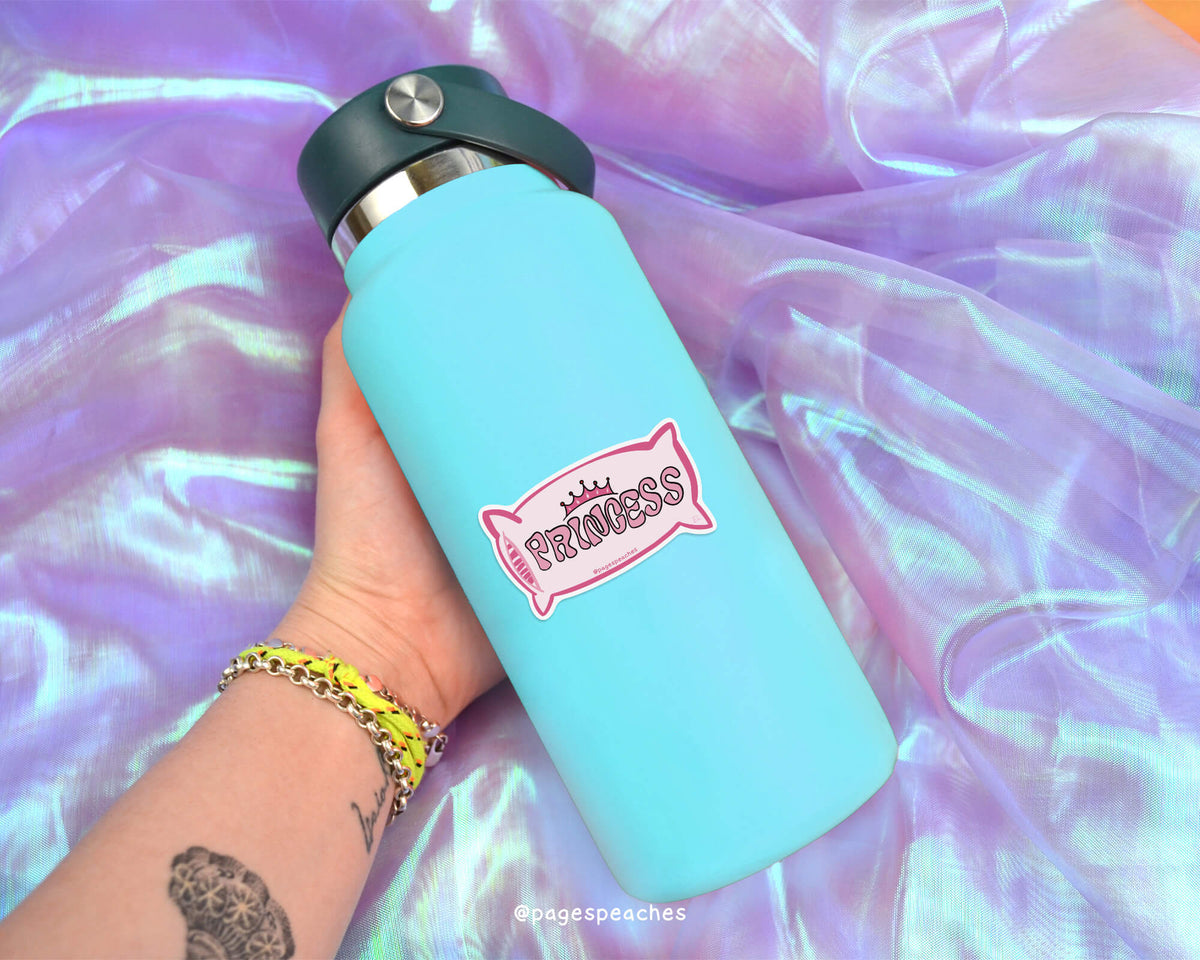 A person&#39;s hand holding a blue water bottle with a pink princess sticker on it, against a purple fabric background.