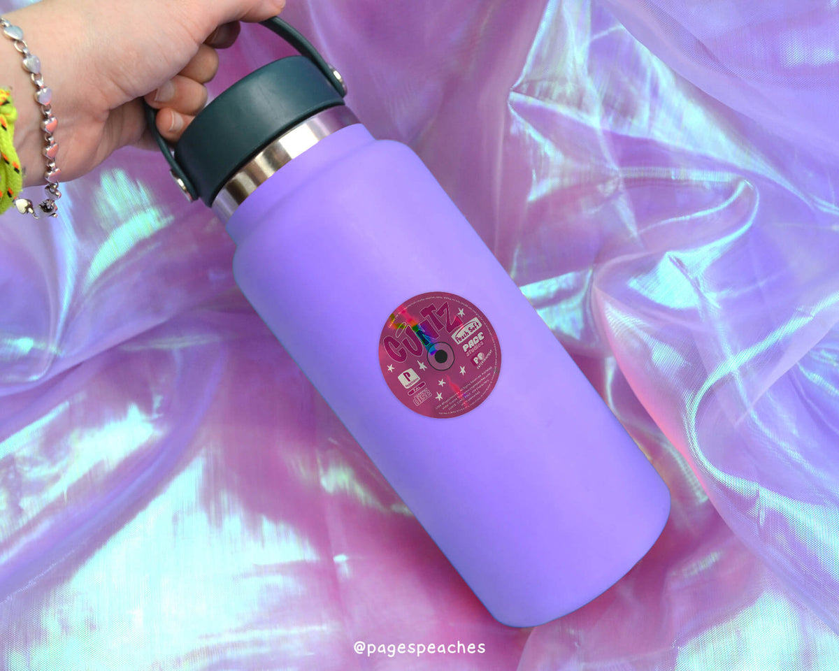 A purple water bottle with a sticker on it, resting on a pink fabric background.
