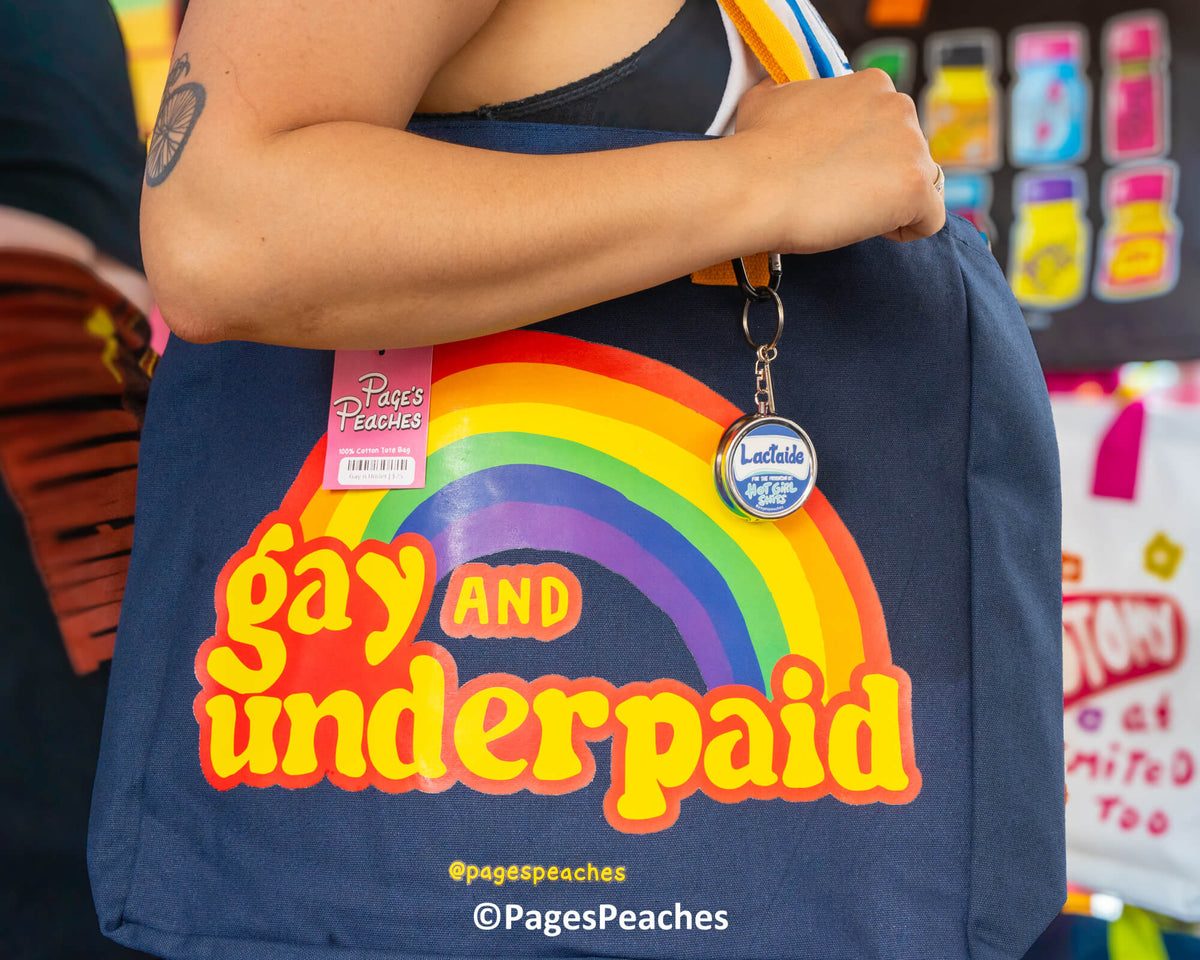 A person is holding a blue tote bag with a rainbow graphic and the text &quot;Gay and underpaid&quot; printed on it.