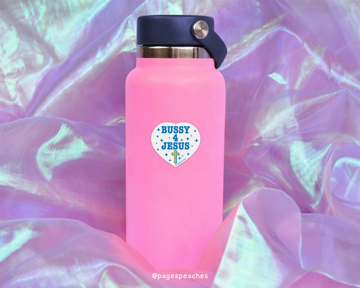 A pink water bottle with a black cap and a sticker that says &quot;BUSY JESUS&quot; on it, placed on a purple fabric background.