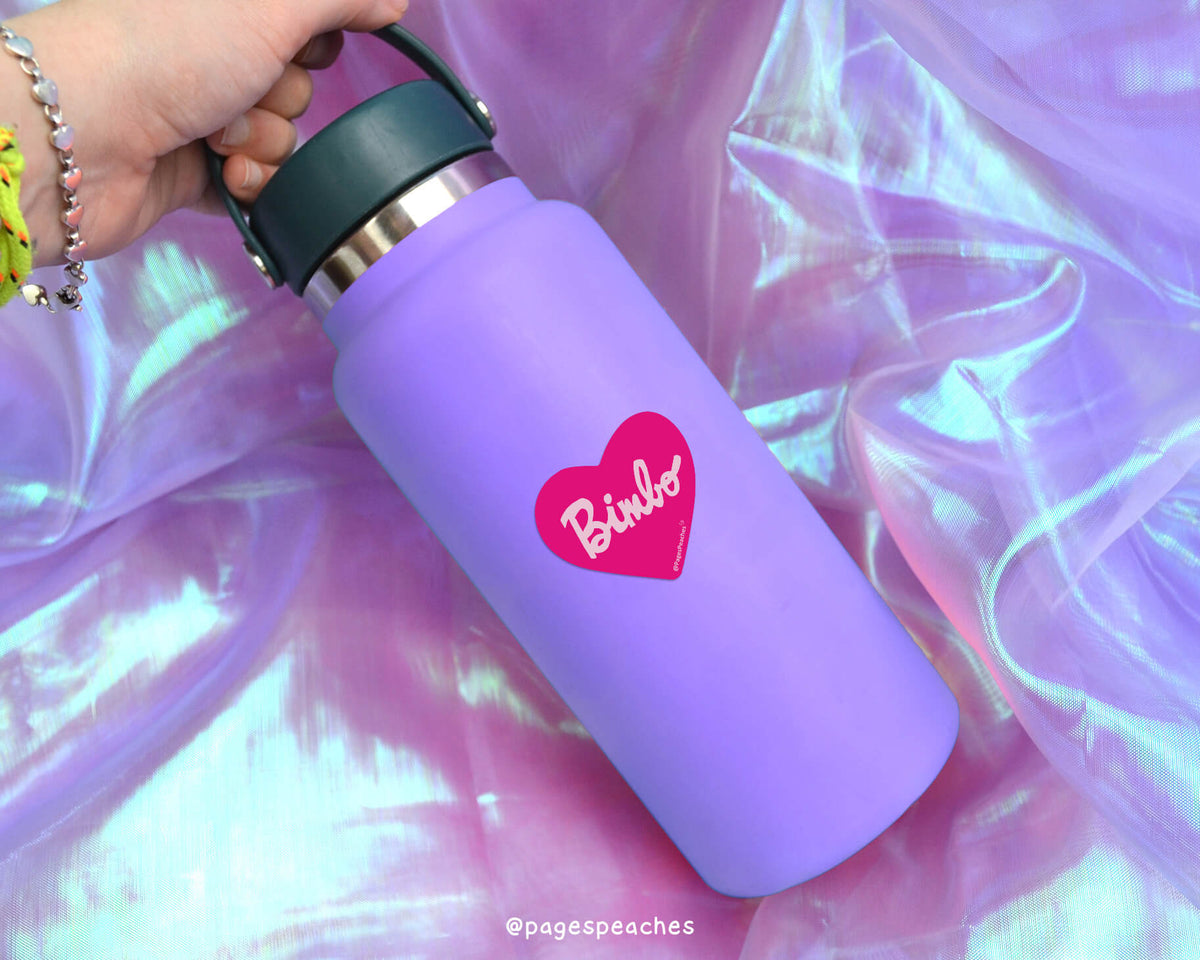 A purple water bottle with a heart-shaped sticker on it, held in a hand against a purple fabric background.