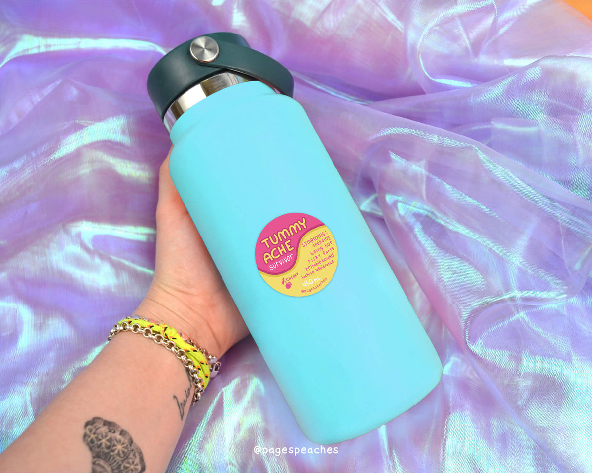 A person&#39;s hand holding a blue water bottle with a pink and yellow label that says &quot;TUMMY ACHE&quot;.