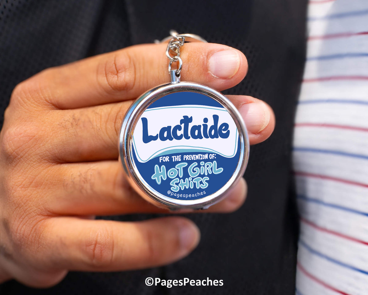 A person is holding a keychain with a circular design featuring the text &quot;Lactaid&quot; and &quot;For the prevention of hot girl shitss&quot; along with a graphic of a hot girl.