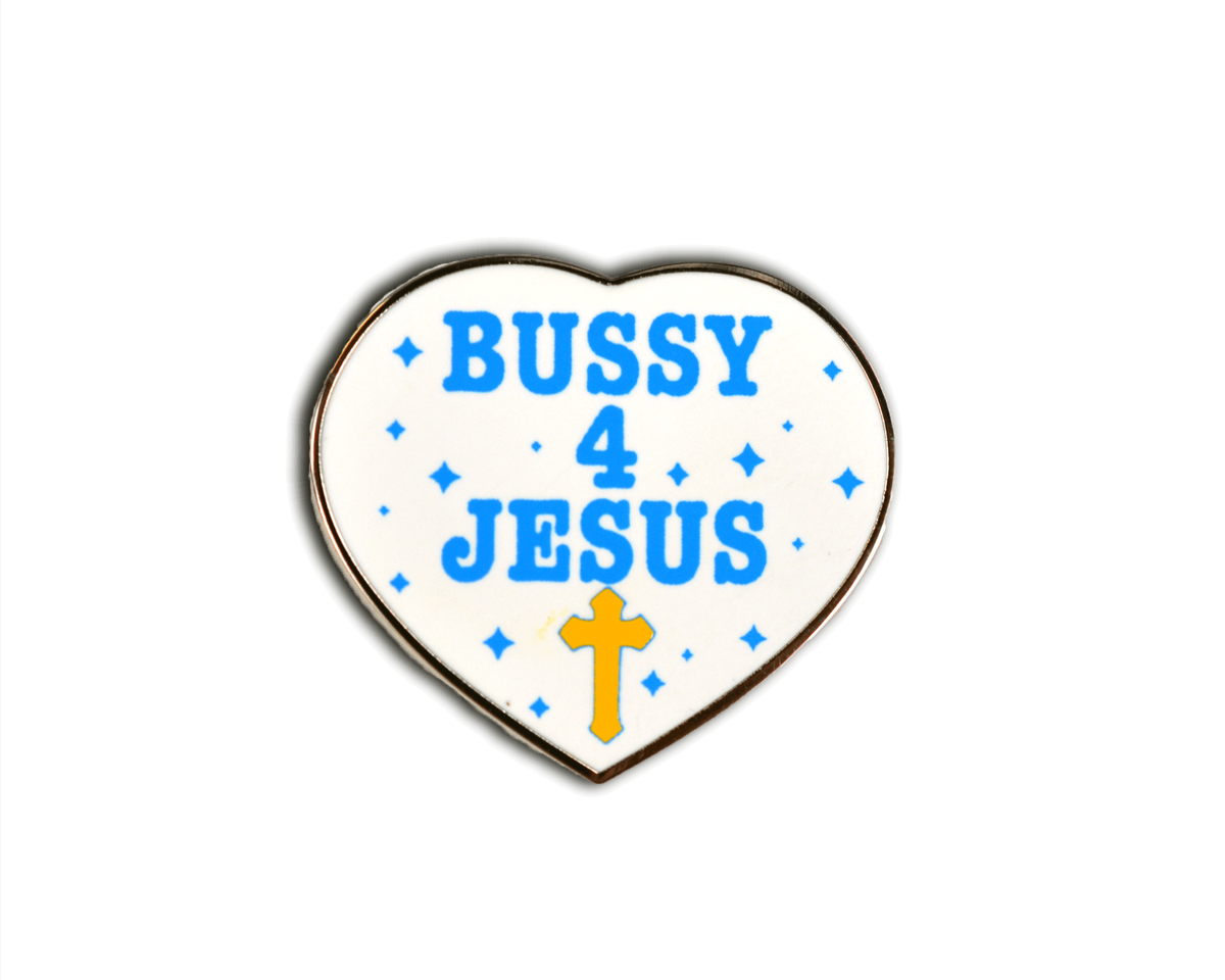A heart-shaped enamel pin with a white face, blue text reading 'BUSSY 4 JESUS', small blue star accents, and a yellow cross at the bottom, shown on a black background.