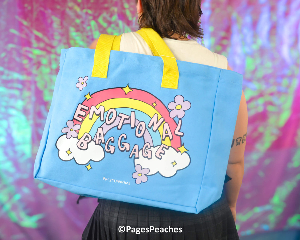 Person carrying a blue tote bag with a rainbow and cloud design, text &#39;Emotional Baggage&#39;, and yellow straps against a colorful background.