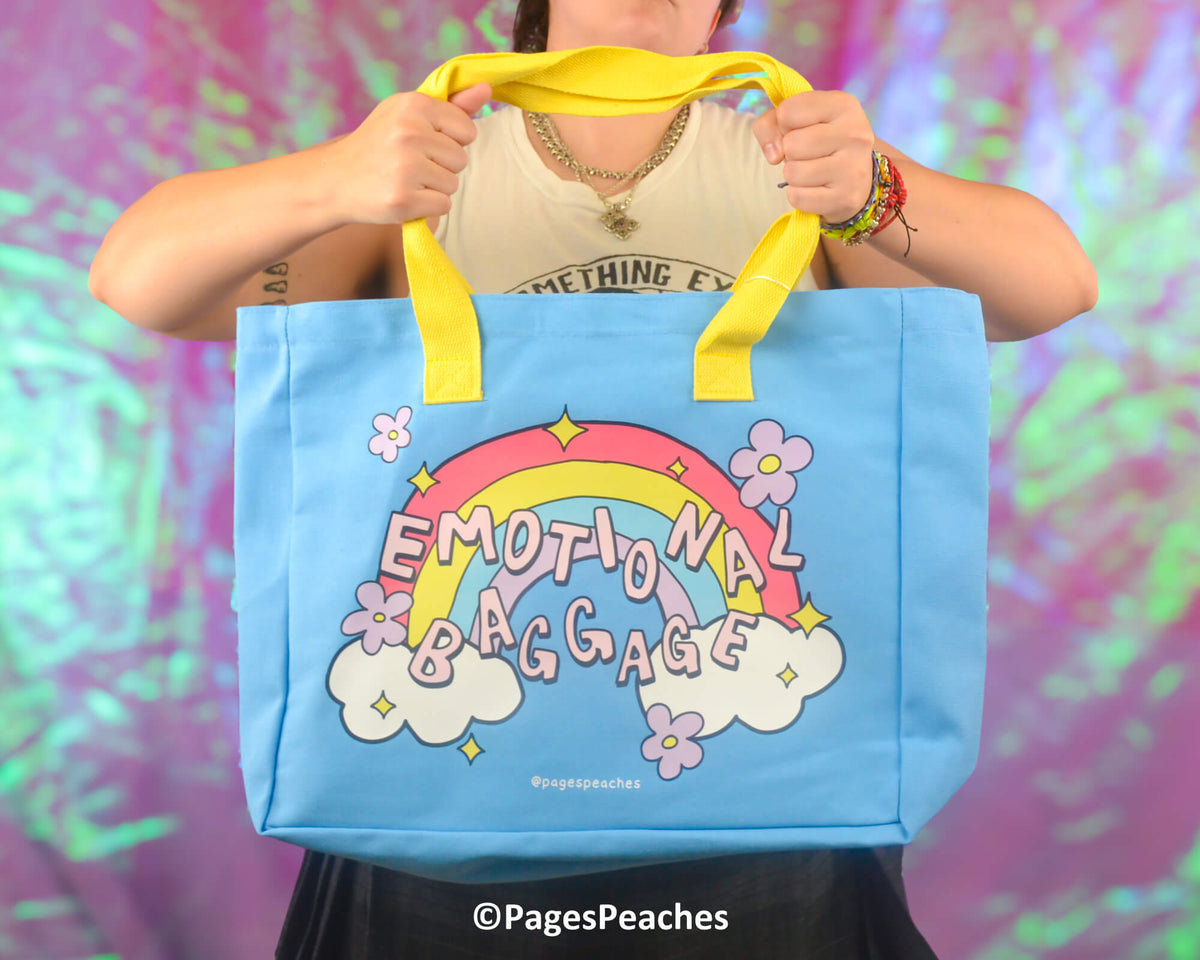Person holding a blue tote bag with a rainbow design and &#39;Emotional Baggage&#39; text against a colorful background.