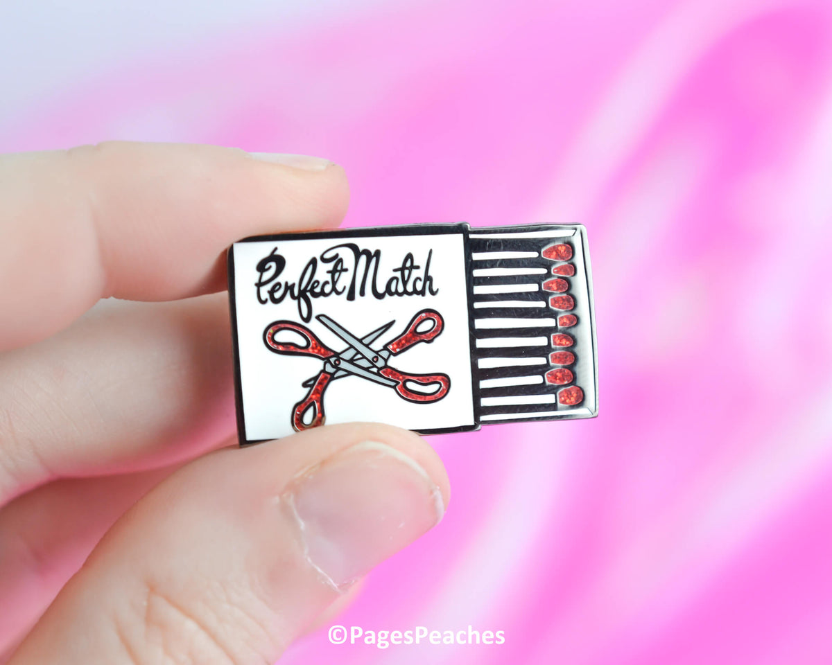 Pin with matchbox design and scissors held by a hand against a pink background
