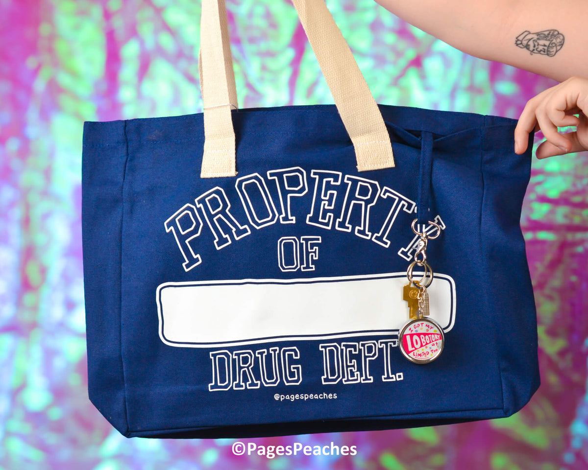 Blue tote bag with &#39;Property of Drug Dept&#39; text and keychain, held by a person against a colorful background.