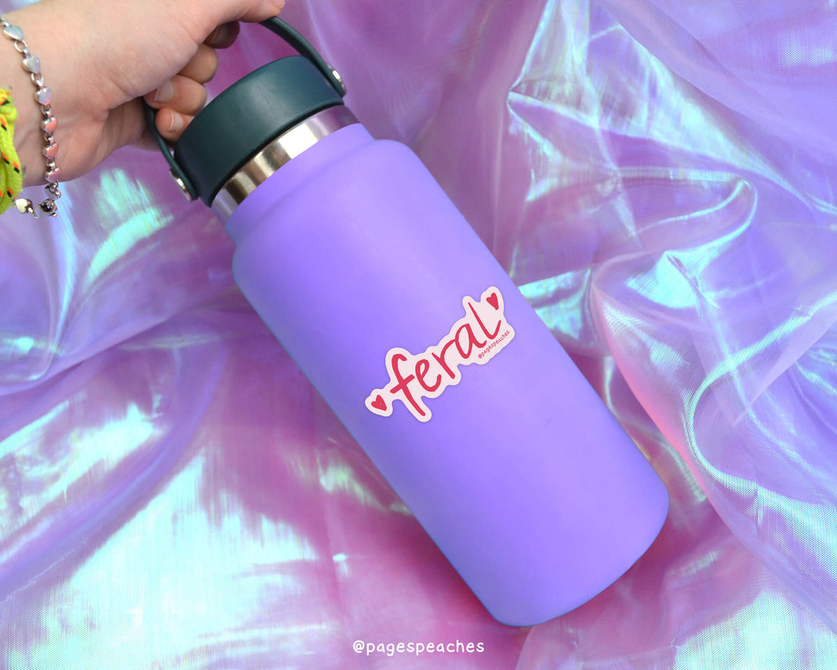 A purple water bottle with a green lid is being held by a person&#39;s hand, with a pink and red sticker on the bottle that says &#39;feral&#39;.