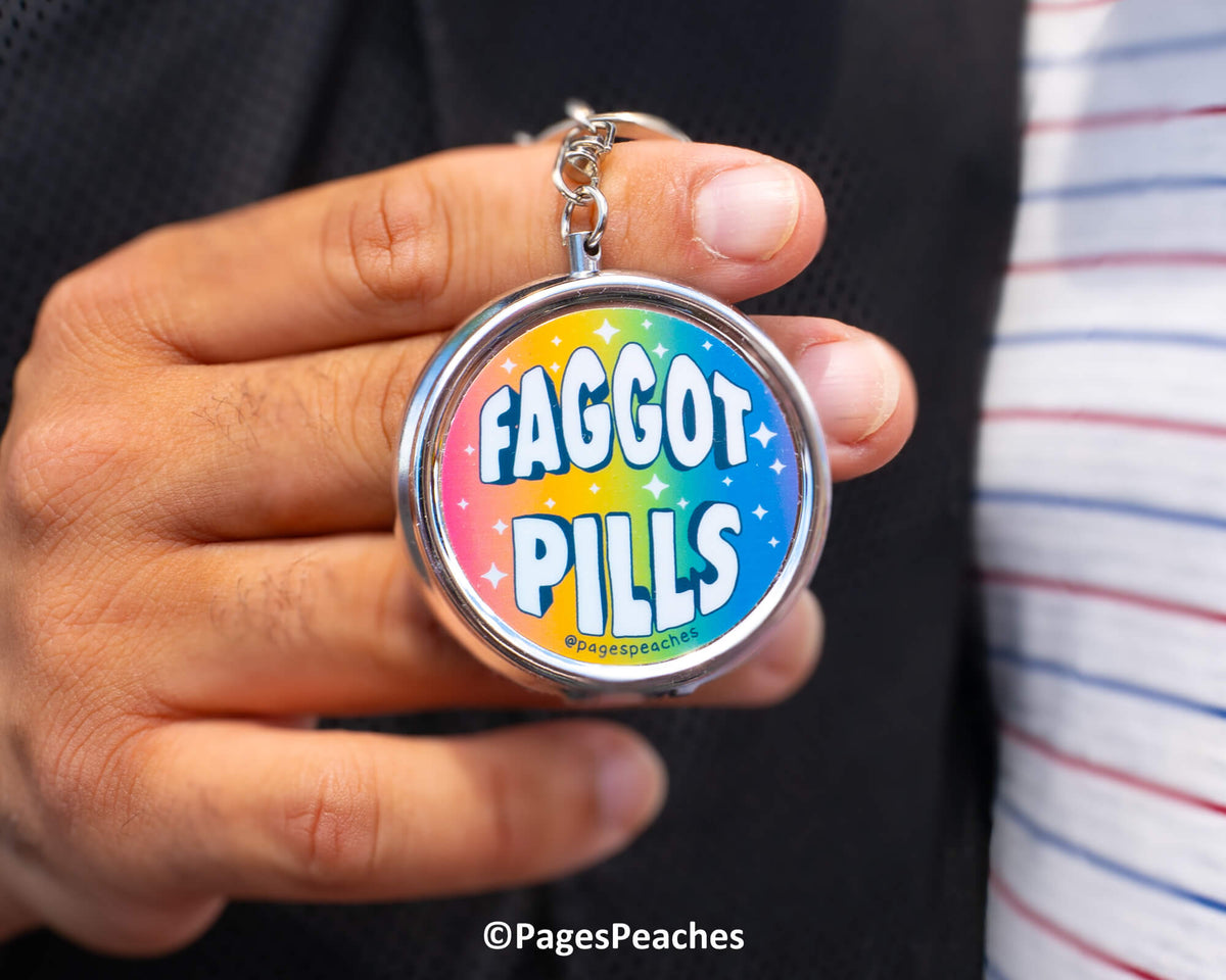 A person is holding a keychain with the text &quot;FAGGOT PILLS&quot; and a rainbow-colored design.