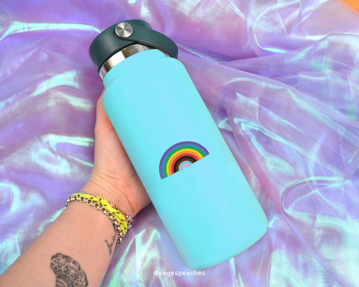 A person&#39;s hand holding a blue water bottle with a rainbow sticker on it, against a purple fabric background.