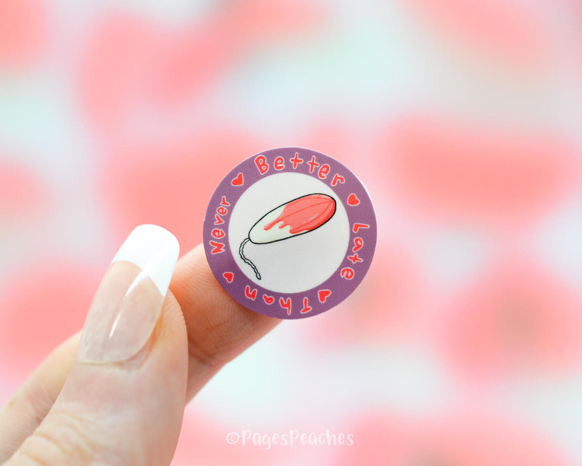 Small Circle Sticker with a bloody tampon in the middle and the outside saying better late than never stuck on a finger
