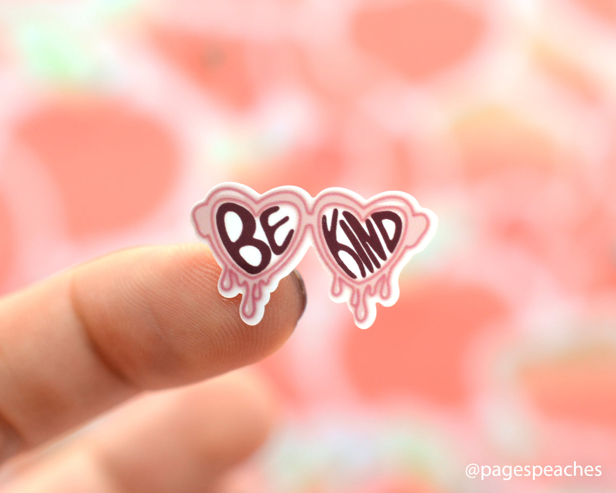 Small melting heart glasses sticker that says be kind stuck on a finger
