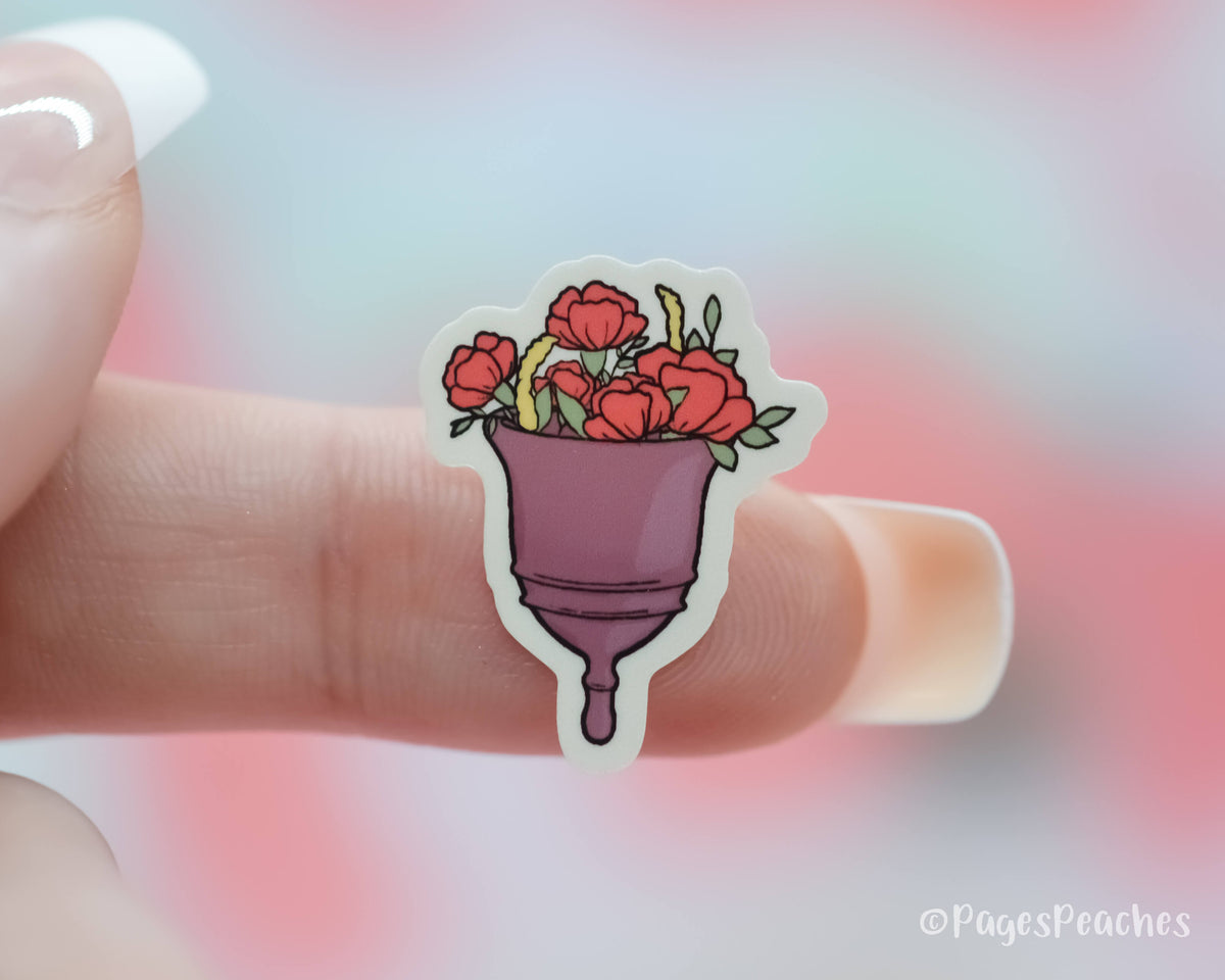 Small Sticker of a purple menstrual cup that has flowers in it stuck to a finger